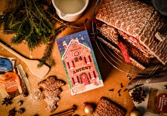 Das Foto zeigt den Gewürzadventkalender mit dem Lebkuchenhaus vorne drauf, der vor einem echten Lebkuchenhaus liegt. Drum herum ist Lebkuchen, weihnachtliche Gewürze und 2 Päckchen aus dem Gewürzadventkalender. Das Foto ist von oben gemacht. | © SONNENTOR