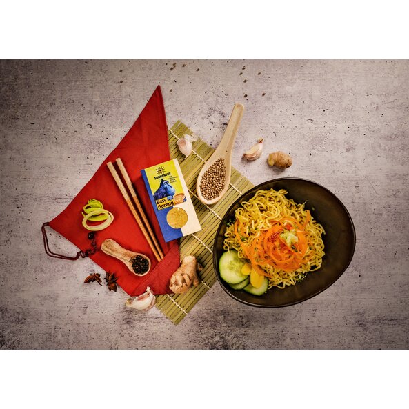 Easy Goreng spice arranged in Asian-stylemix with chopsticks, bamboo mat and ingredients such as garlic and ginger, and a bowl of fried noodles and vegetables on a grey background. 