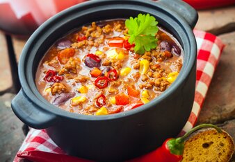 Auf dem Foto ist ein schwarzer Topf mit Chili con Carne auf einem rot kariertem Küchentuch zu sehen. Daneben sind Paprika und Brot zu sehen. | © SONNENTOR