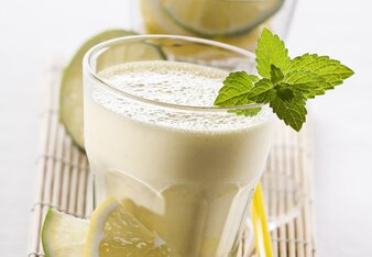 Auf dem Foto ist ein Glas mit Limoncello-Shake zu sehen. Davor sieht man Limetten und Zitronenscheiben. Das Glas ist mit Minze garniert. | © SONNENTOR