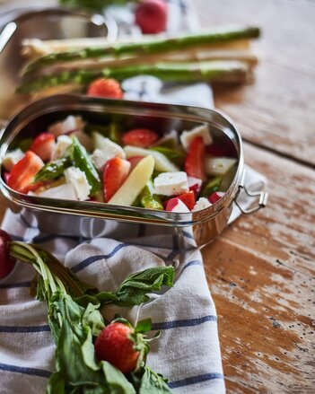 Foto von einer Dose mit buntem Spargelsalat und Feta. Rundherum sind Gemüse und Obst zu sehen. | © SONNENTOR