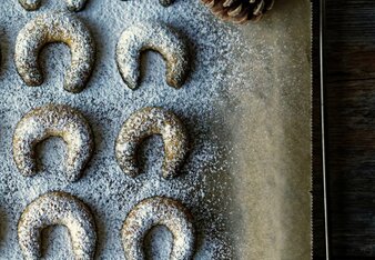 Auf dem Foto sieht man Vanille Kurkuma Kipferl die mit Staubzucker bestreut sind. Daneben sind Zapfen und ein Stern zu sehen. | © SONNENTOR