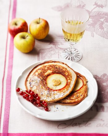 Auf dem Foto ist ein weißes Teller mit Crêpes auf einem rosa Tischtuch zu sehen. Dahinter sind Äpfel zu sehen. | © SONNENTOR