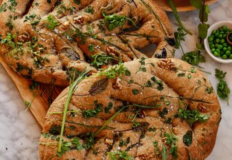 Zwei Stück Brot-Gebäck mit regelmäßigen Einschnitten, belegt mit Walnüssen und Wildkräutern, die fast aussehen, wie ein Zopf, liegen auf einem Holzbrett. | © SONNENTOR