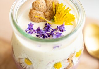 Die Topfen Creme ist in einem bauchigen Glas, dekoriert mit einem Durch die Blume Keks und Blüten von Wildkräutern. Durch das Glas sieht man außerdem Gänseblümchen, die in der Creme in einer Reihe trapiert sind. | © SONNENTOR