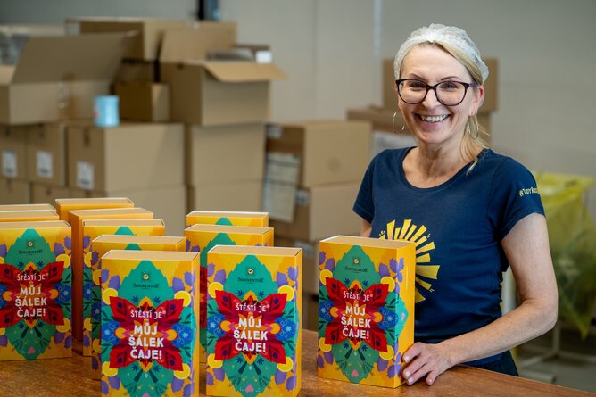Na fotografii je usmívající se spolupracovnice SONNENTORU vedle stolu, na kterém jsou vyskládané dárkové kazety s biočaji SONNENTOR. Na kazetě je vidět nápis Štěstí je můj šálek čaje. | © SONNENTOR