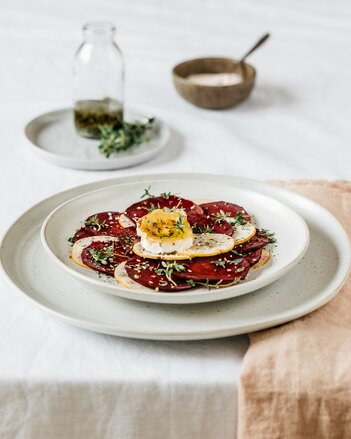 Auf dem Foto sieht man ein Teller mit rotem Beete Carpaccio. Darauf liegt Ziegenkäse. Im Hintergrund sieht man Gewürze und Kräuter. | © SONNENTOR