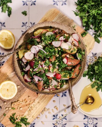 Auf dem Foto ist eine Schüssel mit Kartoffel-Radieschen Salat zu sehen. Die bunte Bowl steht auf einem Holzbrett. Daneben sind Pinienkerne zu sehen. | © SONNENTOR