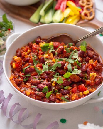 Das Chili Sin Carne besteht aus Bohnen, Mais und Sauce und ist mit grünen Blättern dekoriert in einer weißen Schüssel mit einem Löffel darin.  | © SONNENTOR