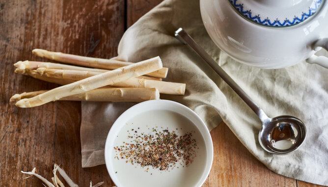 Auf dem Foto ist eine Schüssel mit weißer spargelcremesuppe zu sehen. Darauf sind verschiedene Kräuter gestreut. Daneben sieht man ganzen Spargel. | © SONNENTOR