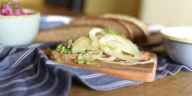 Auf dem Foto sieht man ein Holzbrett mit einer Scheibe Brot darauf. Das Brot ist mit Apfel, Erbsen und Fenchel belegt. | © SONNENTOR